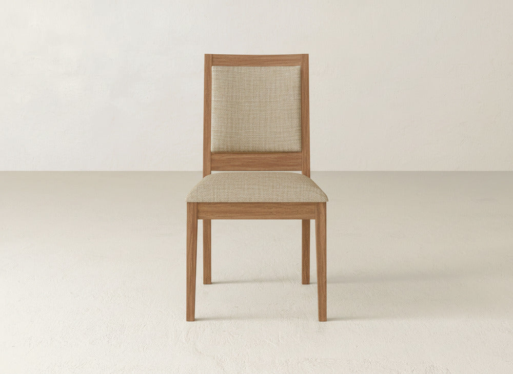 A James+James Gina Side Dining Chair, featuring maple wood and a beige upholstered seat and backrest, sits on a light floor against an off-white wall.