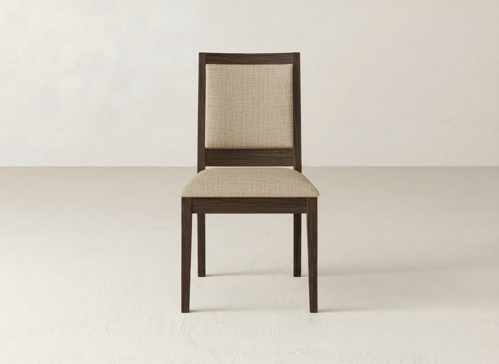 The Gina Side Dining Chair by James+James, with a beige upholstered seat and backrest, showcases clean lines and dark maple wood in a simple modern design against a plain, light-toned wall and floor.