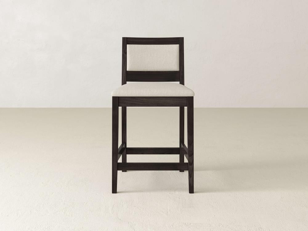 A tall wooden chair with a high backrest and beige cushioned seat, echoing the Gina Side Stool's modern design by James & James, stands centered. Crafted from maple wood, it adorns a plain, light-colored floor against an off-white wall.