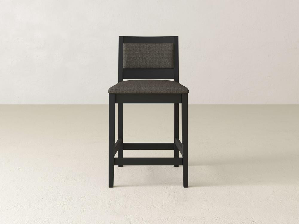 The Gina Side Stool by James & James features a modern design with a sleek black frame and dark cushioned seat and backrest, beautifully contrasting against a plain white wall and light beige floor.