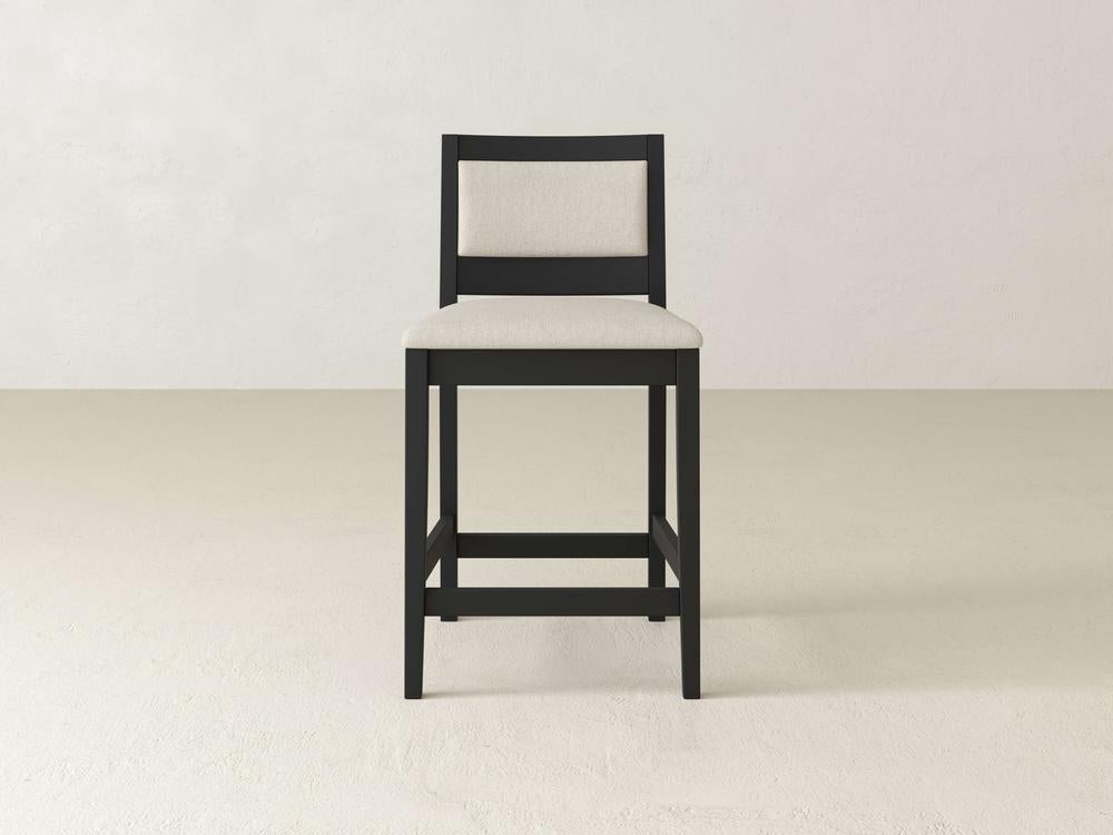 The Gina Side Stool by James & James, featuring black maple wood and tailored beige upholstery, elegantly complements a light-colored floor against a plain white wall with its simple modern design.