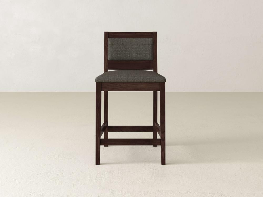 The Gina Side Stool by James & James is a modern wooden bar stool crafted from maple with a dark brown finish. It features a cushioned backrest and padded seat in dark gray fabric, set against a plain white background.