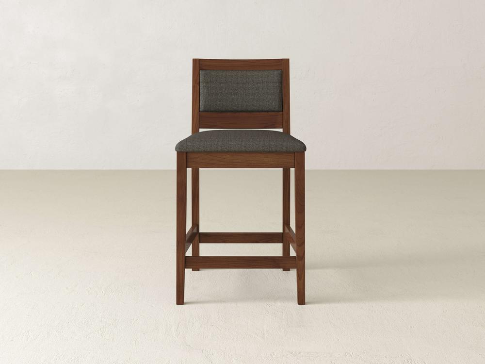 The Gina Side Stool by James & James features a dark green padded seat cushion and backrest, standing against a plain light beige wall and floor. Its minimalist, modern design highlights the natural grain of maple wood.