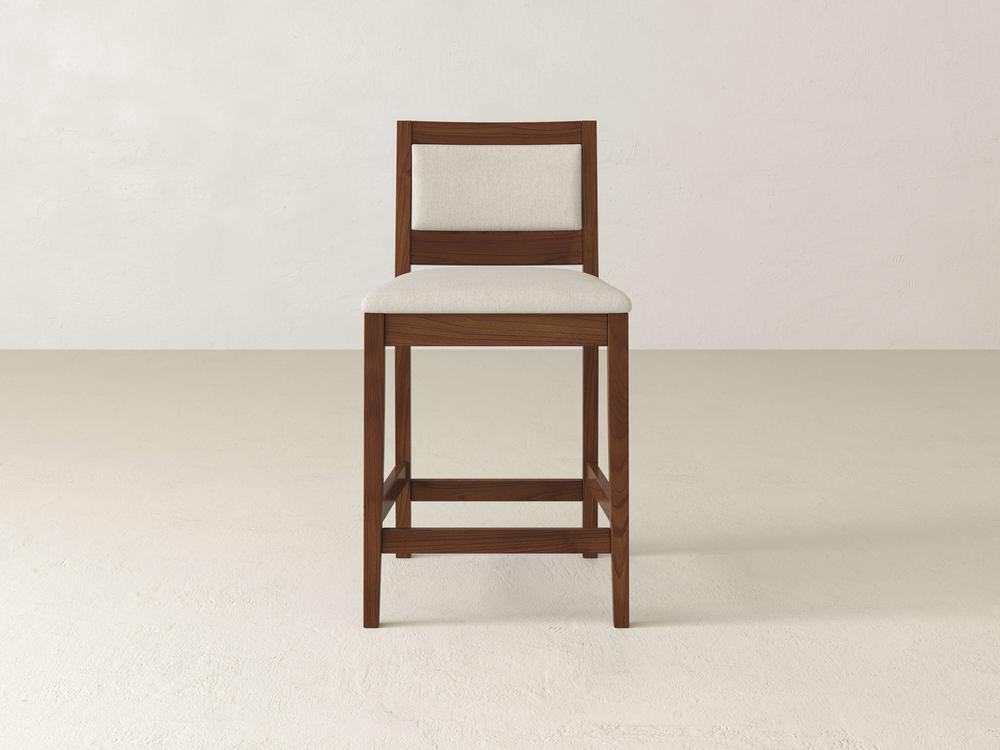 The Gina Side Stool by James & James, crafted from Maple wood, features a modern silhouette with a beige padded seat and backrest. Its simple design includes a square frame and footrest, and it is pictured against a plain white wall.