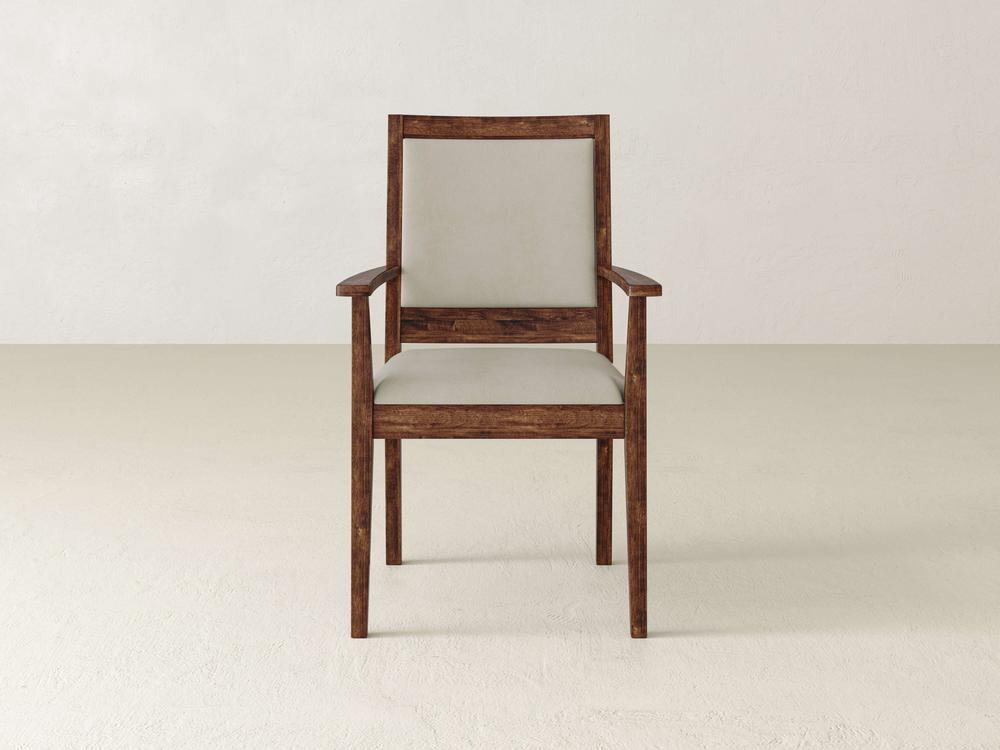 A wooden chair with a beige cushioned seat and backrest is centered against a plain white wall. The chair has armrests and a minimalist design. The floor is light-colored, complementing the simple, elegant aesthetic of the chair.