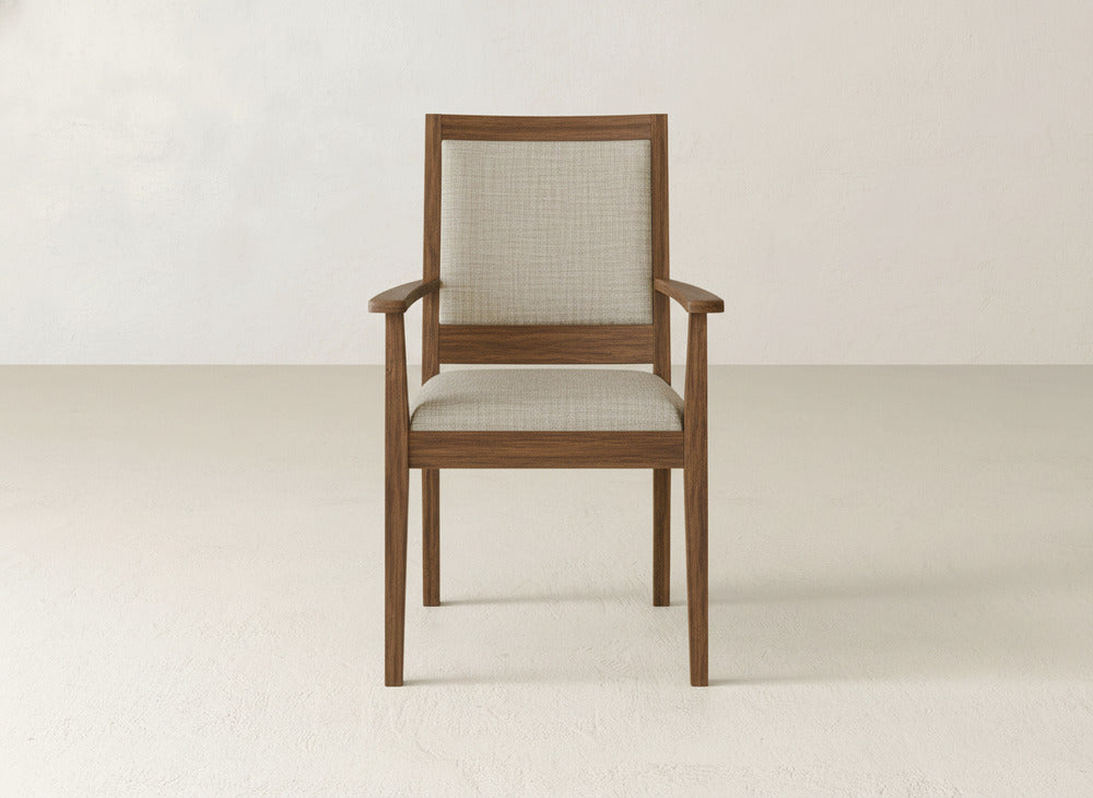 A James+James Gina Arm Dining Chair, crafted from maple with a light beige cushioned seat and backrest, sits in a minimalist room featuring light-colored walls and flooring.