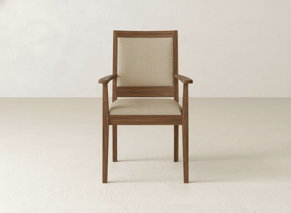 The Gina Arm Dining Chair by James+James features a maple wood frame with beige upholstered seat and backrest, shown in an empty room with a plain light-colored wall and floor.