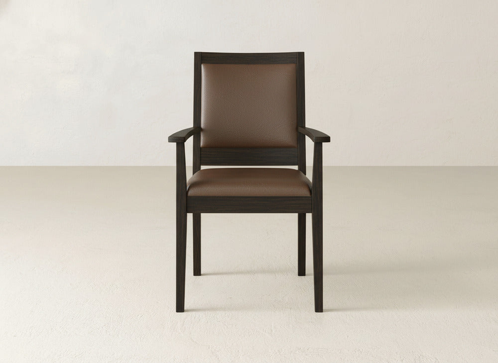 The Gina Arm Dining Chair by James+James features a padded brown leather seat and backrest, crafted from maple wood, set on a light floor with an off-white wall backdrop.
