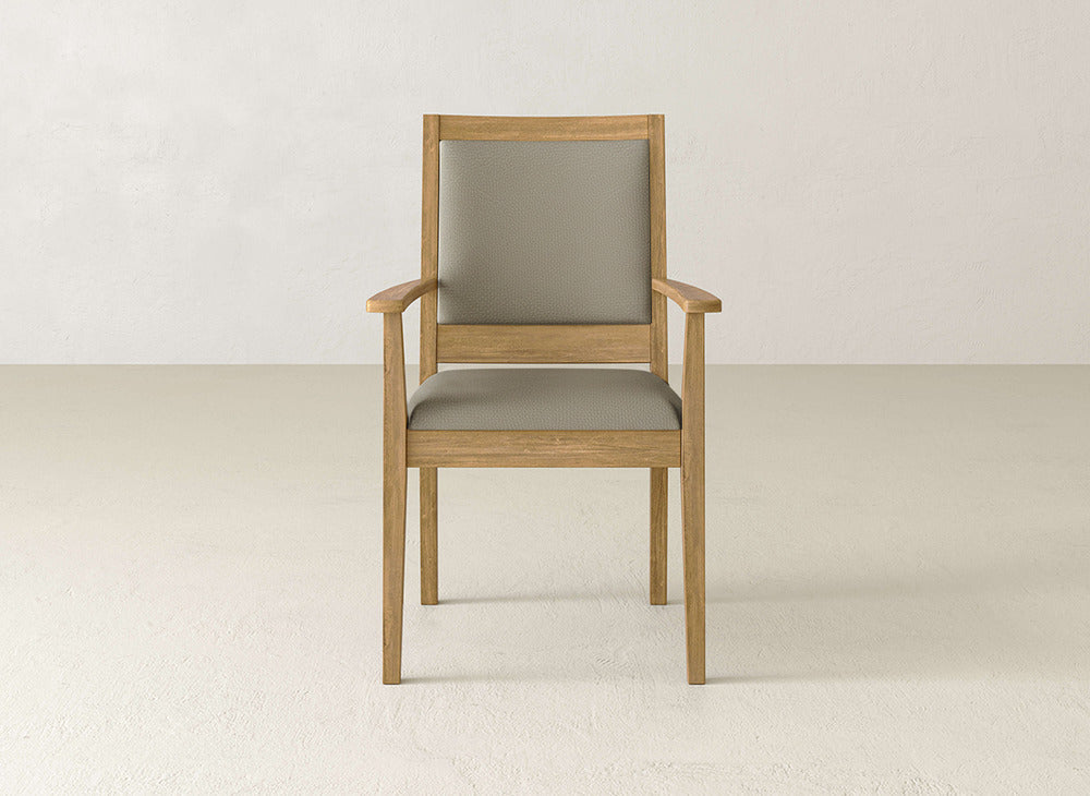 The James+James Gina Arm Dining Chair features a maple wood frame in light brown and a light gray upholstered seat and backrest, shown on a neutral floor with a plain, light-colored wall in the background.
