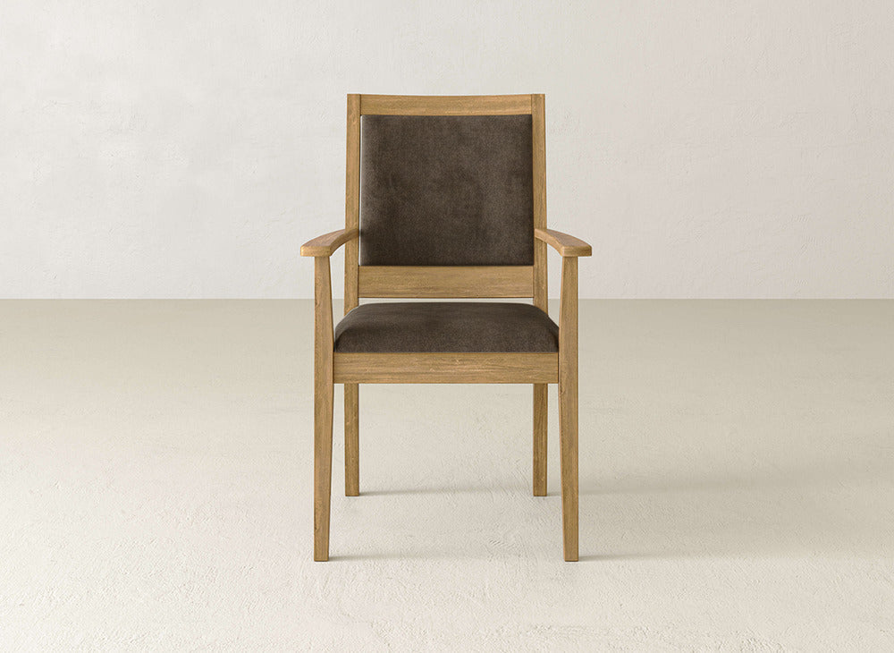 The Gina Arm Dining Chair by James+James features maple wood armrests and a brown cushioned seat and backrest, shown on a light floor against an off-white wall.