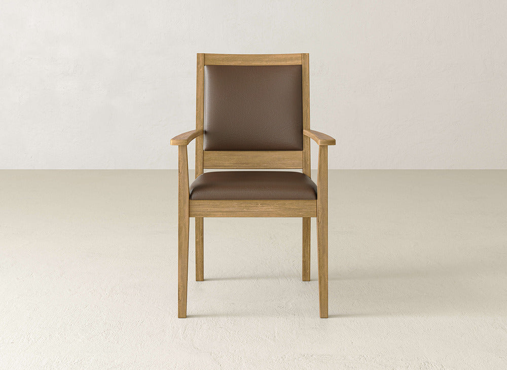 The Gina Arm Dining Chair by James+James is a modern maple wood chair with armrests and a brown cushioned seat and backrest, placed on a light floor against an off-white wall.