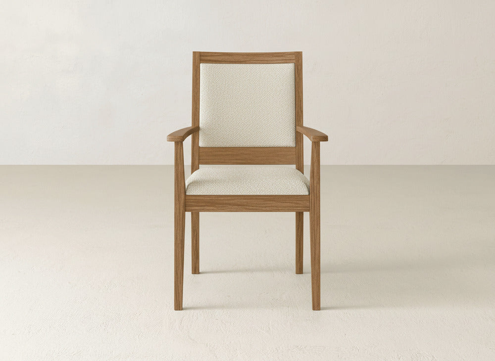 The Gina Arm Dining Chair by James+James features a maple wood frame and light beige upholstered seat and backrest, shown against a plain, light-toned wall with a matching floor.