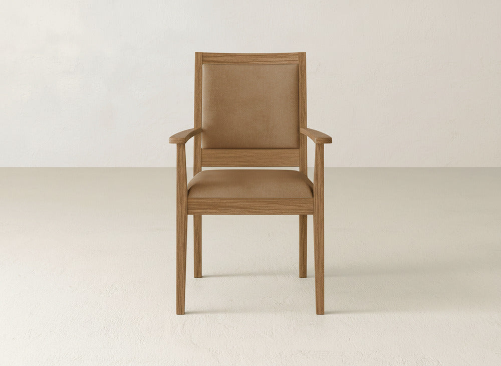 The Gina Arm Dining Chair by James+James showcases a maple wood frame with a brown cushioned seat and backrest, elegantly placed in an empty room with off-white walls and a light floor—a stylish, modern dining chair for any setting.