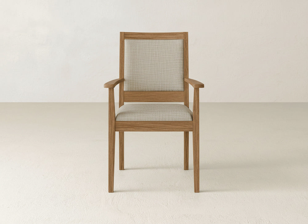 The Gina Arm Dining Chair by James+James features maple wood, armrests, and a light beige upholstered seat and backrest, shown against a simple, light-toned wall and floor.