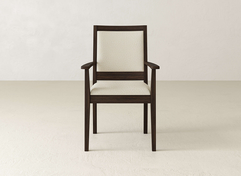 The James+James Gina Arm Dining Chair features a dark brown maple frame and light beige upholstery, bringing contemporary elegance to your dining space when set on a light floor against an off-white wall.