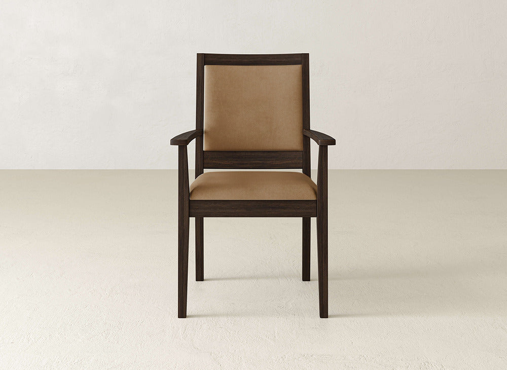 The James+James Gina Arm Dining Chair, crafted from maple with brown cushioned seating and armrests, is elegantly displayed in a minimal, light-toned room featuring plain walls and flooring.