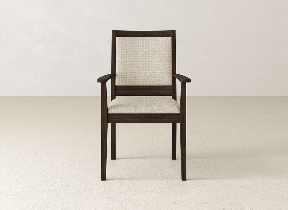 The James+James Gina Arm Dining Chair is a contemporary dining chair with a dark brown maple wood frame and light beige upholstered seat and backrest, shown on a light floor against an off-white wall.