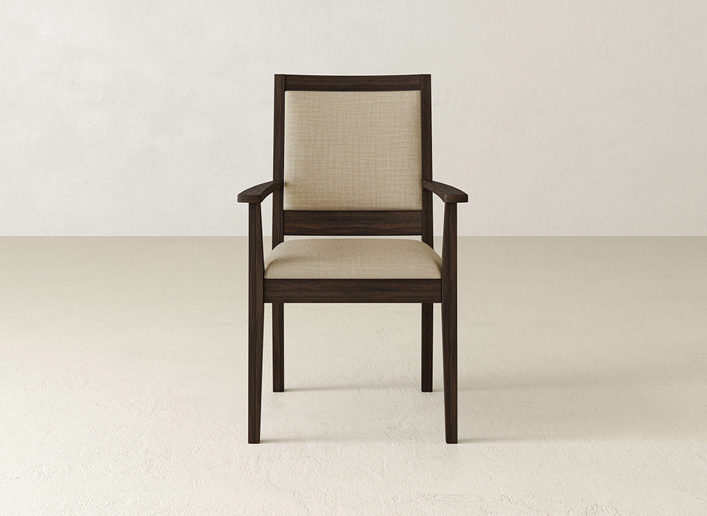 The James+James Gina Arm Dining Chair, crafted from maple with armrests and a beige upholstered seat and back, is showcased at the heart of a minimalist, light-toned room.