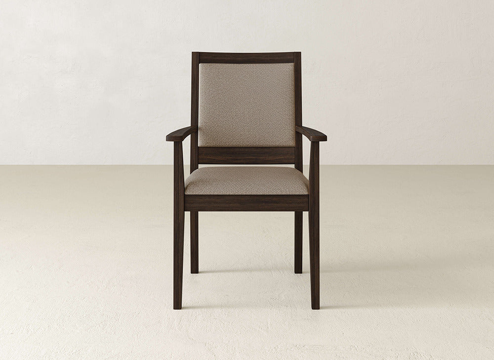 The Gina Arm Dining Chair by James+James features a contemporary dark brown maple wood frame, upholstered backrest, and beige cushioned seat. It’s shown on a light floor with an off-white wall background.