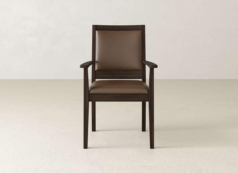 The James+James Gina Arm Dining Chair is a modern maple dining chair with armrests and brown cushioned seat and back, displayed on a light floor with a plain white wall backdrop.