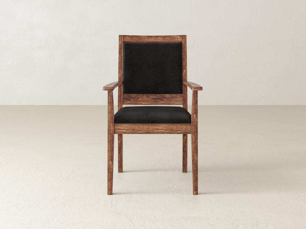 A wooden chair with a dark cushioned seat and backrest is centered against a plain, light-colored wall and floor. The chair has armrests and a simple, traditional design.