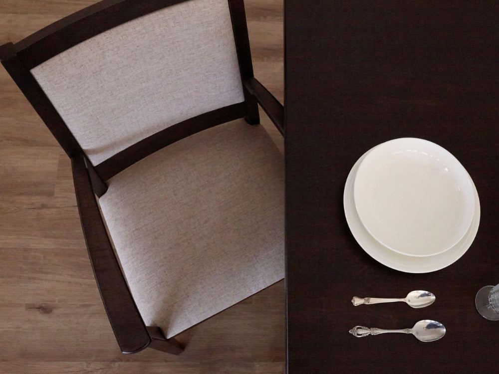 A top-down view of a James+James Sand - Linen Swatch dining chair with a light cushion beside a dark table set with stacked white plates, glassware, and cutlery on a wooden floor.