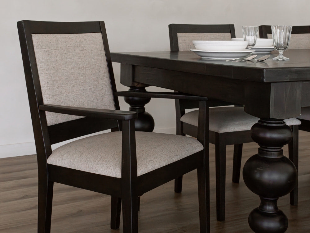 A dark wood dining table with turned legs exudes timeless elegance, set with white dinnerware. Two James+James chairs upholstered in Sand - Linen Swatch fabric add airy softness to the space atop a wooden floor.