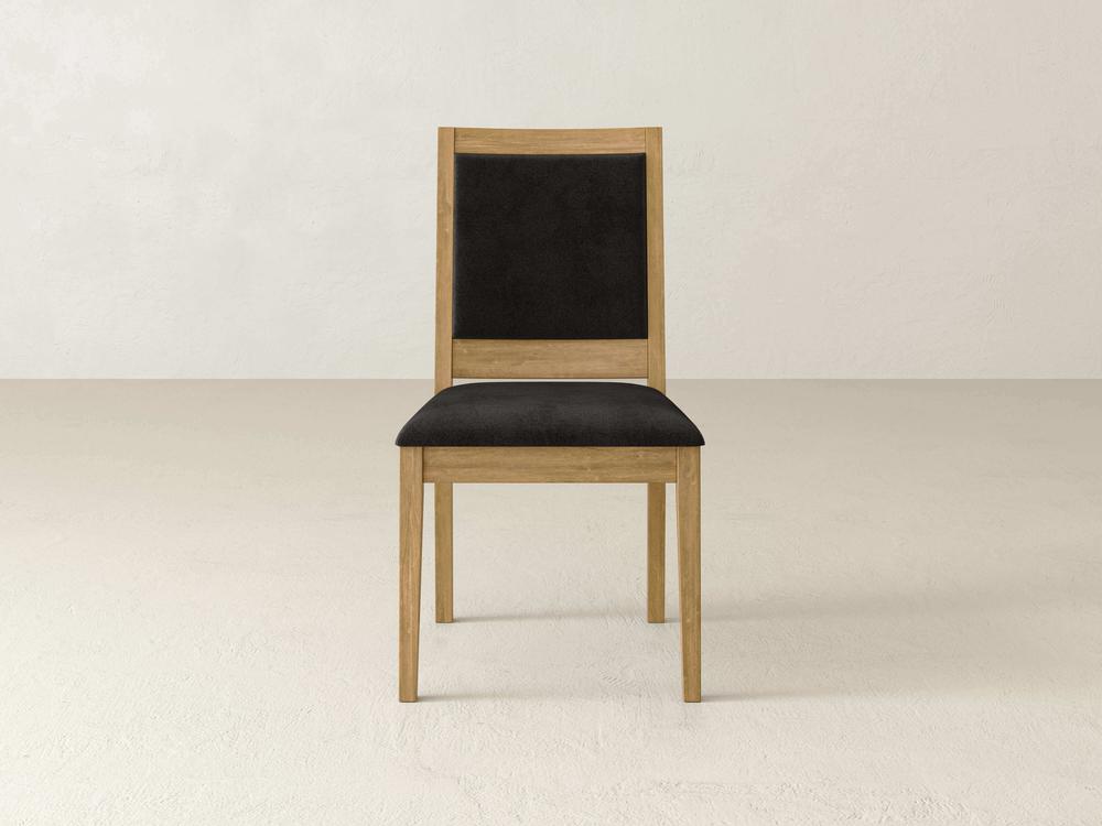 A wooden chair with a black cushioned seat and backrest is centered in a plain room with a light-colored floor and walls. The chair features a simple, modern design with clean lines.