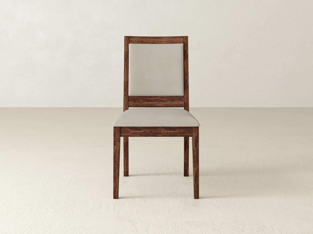 A single wooden chair with light upholstery, positioned on a neutral-colored floor, is centered against a plain, light-colored wall.