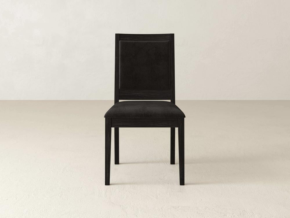 A black wooden chair with a cushioned seat and backrest sits on a beige floor against a plain, light-colored wall. The chair has a simple, elegant design with clean lines.