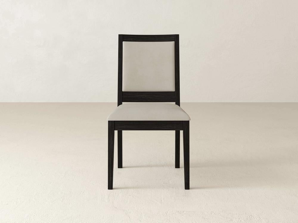 A modern, minimalist chair with a dark wood frame and light beige upholstery is centered in a plain room with a light-colored floor and wall.
