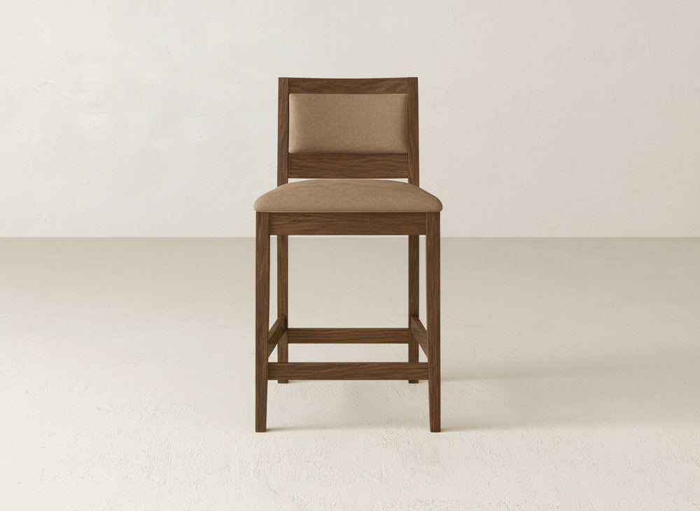 The Gina Side Stool by James & James showcases a modern square backrest and seat in light brown fabric, crafted from maple wood. This wooden stool is set on a plain, light floor with a white wall background.
