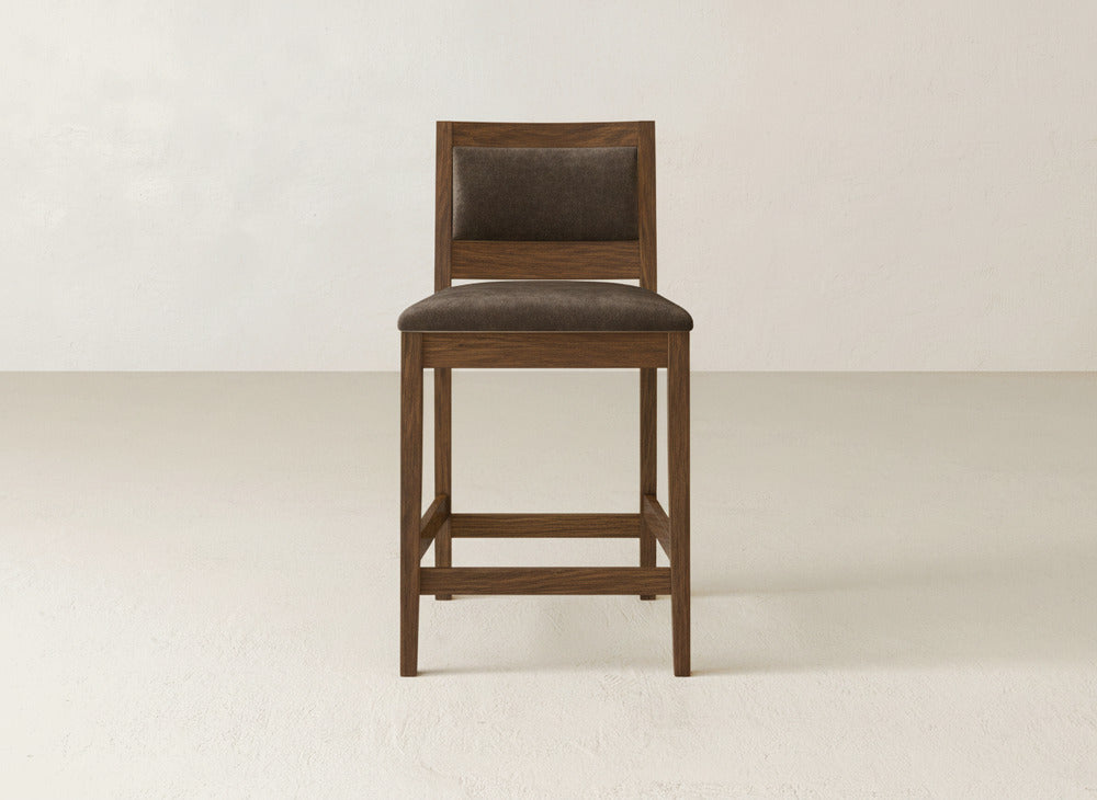 The Gina Side Stool by James & James features a modern design with a dark brown padded seat and backrest, crafted from maple wood, displayed on a light floor against an off-white wall.