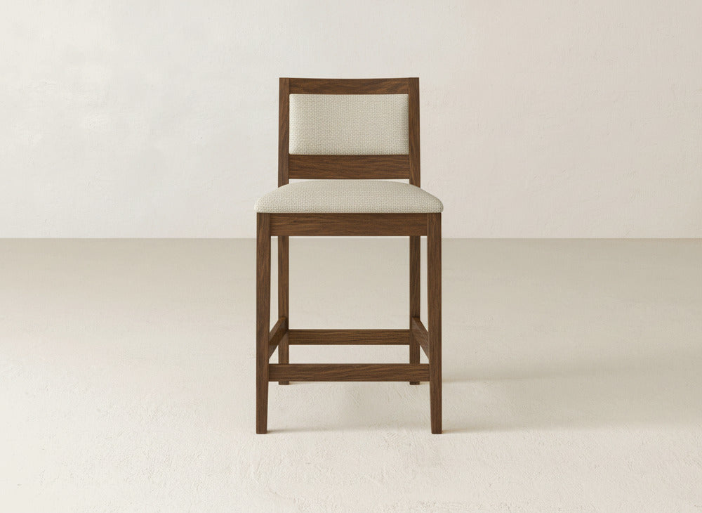 A Gina Side Stool by James & James, featuring a modern maple wood frame with light upholstery on the seat and backrest, sits against a plain light-colored wall and floor.