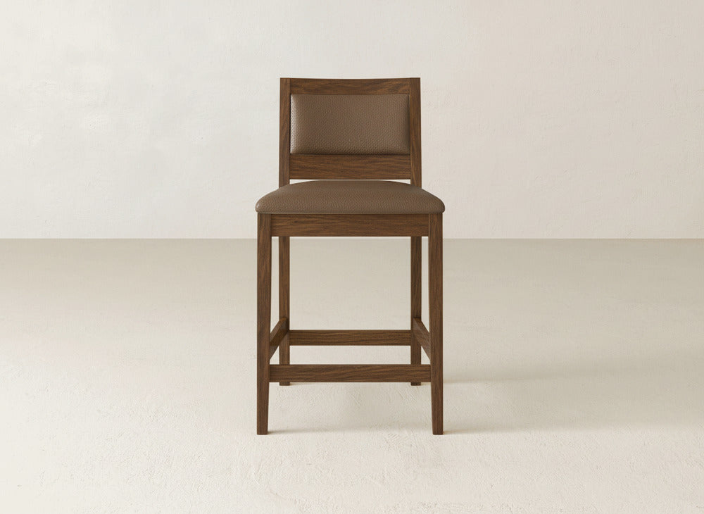 The Gina Side Stool by James & James features a simple, modern design with a padded brown seat and backrest, crafted from maple wood. It stands on a light floor against an off-white wall for a clean, contemporary look.