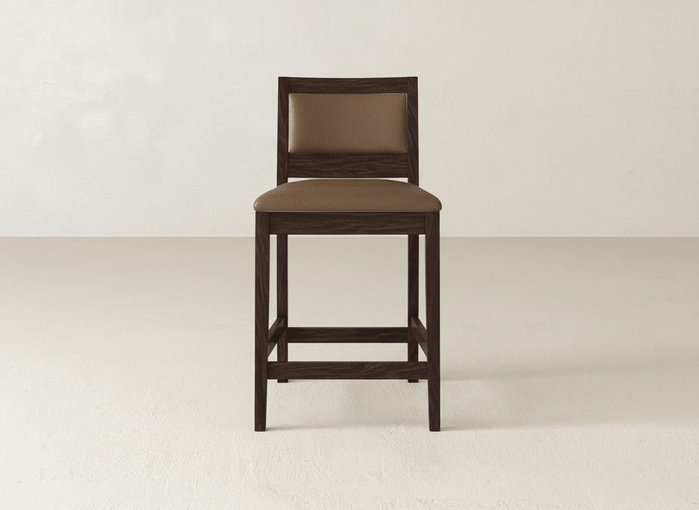 The Gina Side Stool by James & James showcases a modern design with a dark brown maple wood frame and cushioned brown-upholstered seat and backrest, displayed on a light floor against an off-white wall.