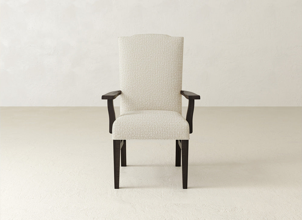 The James+James Hadley Arm Dining Chair features light beige upholstery, a high back, and dark wood legs and arms—an elegant dining chair placed against an off-white wall on a light-colored floor.