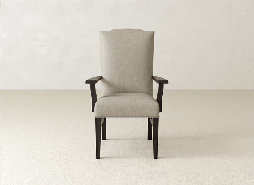 The Hadley Arm Dining Chair by James+James features beige upholstery, a high back, armrests, and dark wooden legs. It stands against a light wall and floor in a minimal, empty room.