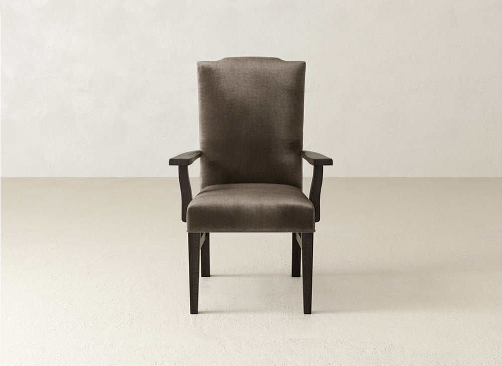 A straight-on view of the James+James Hadley Arm Dining Chair, a brown high-back chair with wooden legs and armrests, set in a minimalist room with a light floor and plain white wall.