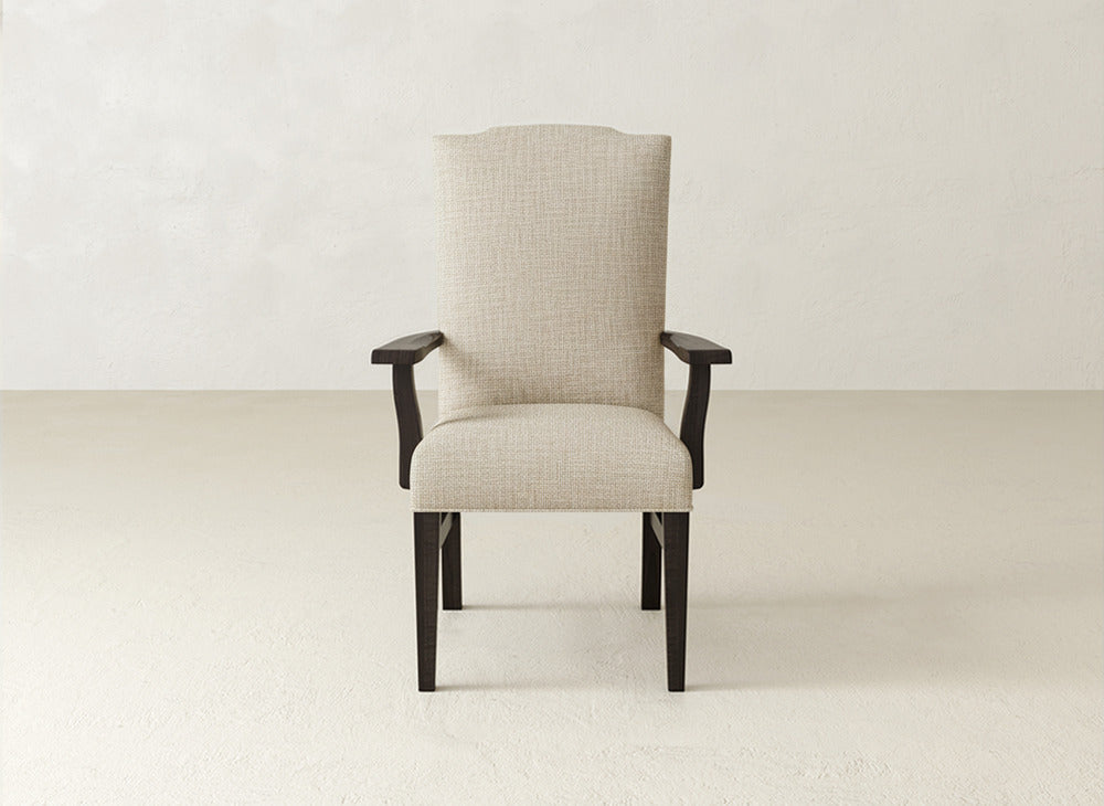 A Hadley Arm Dining Chair by James+James, featuring beige upholstery and dark wooden legs and armrests, is centered in a minimalist room with a light wall and floor.