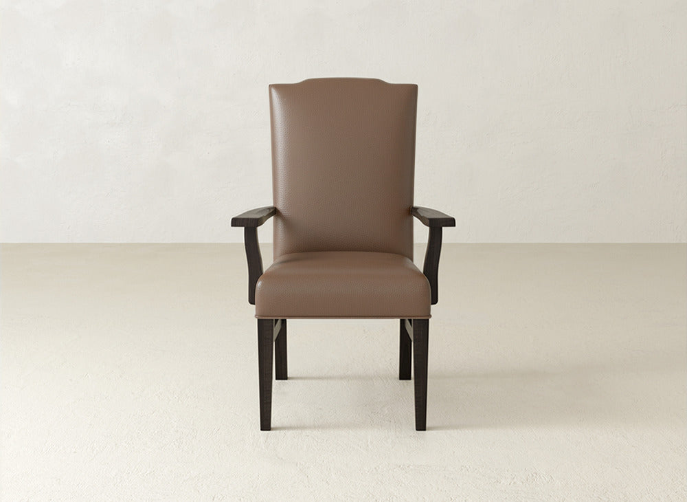 A brown Hadley Arm Dining Chair by James+James, with dark wood legs and arms and an upholstered high back, is set on a light floor against a textured off-white wall.