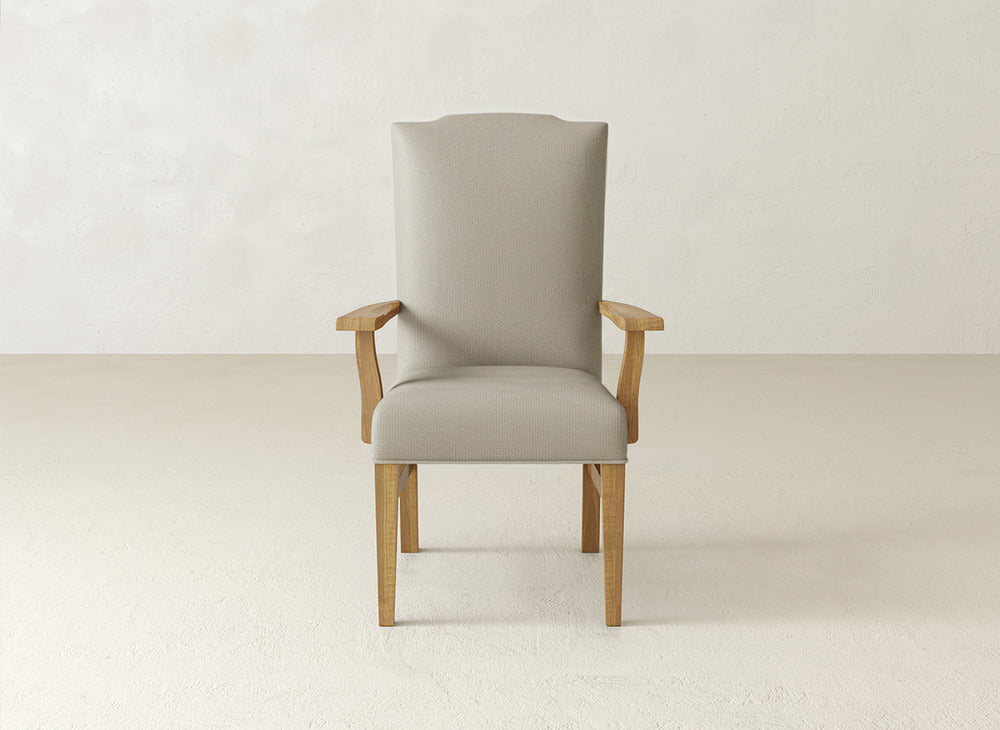 The Hadley Arm Dining Chair by James+James, featuring beige upholstery and wooden arms and legs, is centered in a minimalist room with light-colored walls and flooring.