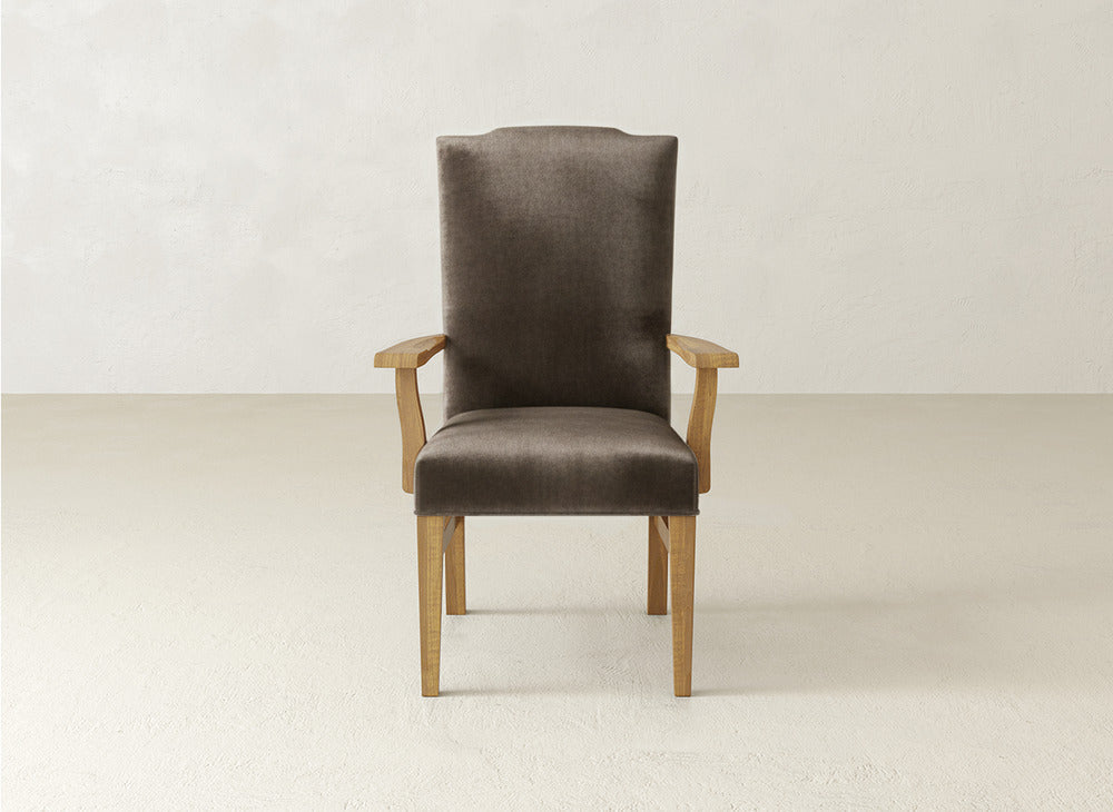 The Hadley Arm Dining Chair by James+James, featuring a high backrest and cushioned seat in dark brown upholstery, is centered against a plain, light-colored wall and floor.