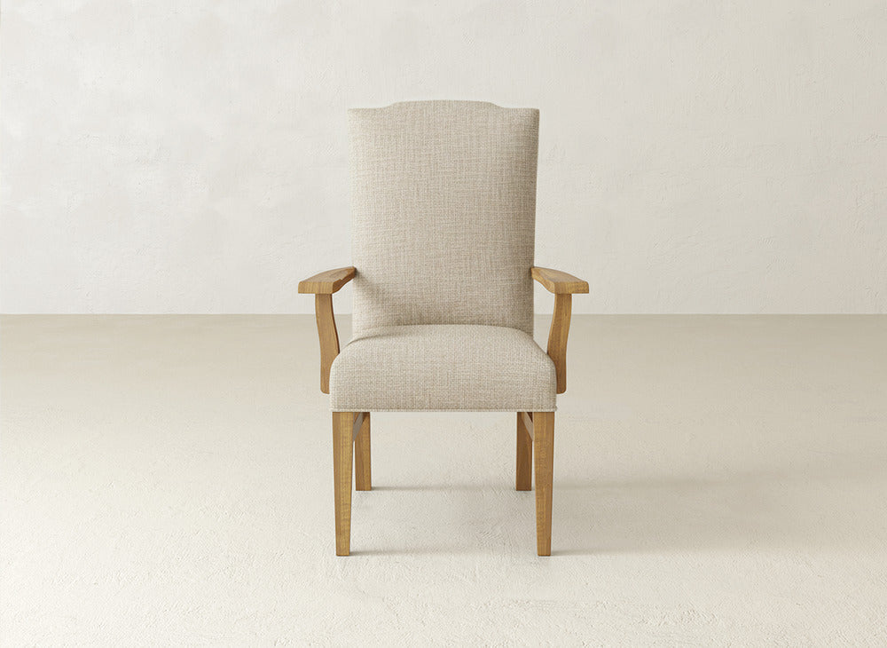 The Hadley Arm Dining Chair by James+James features beige upholstery with wooden legs and arms, set against an off-white wall and floor in a minimalist space, providing the comfort and style of an armchair.
