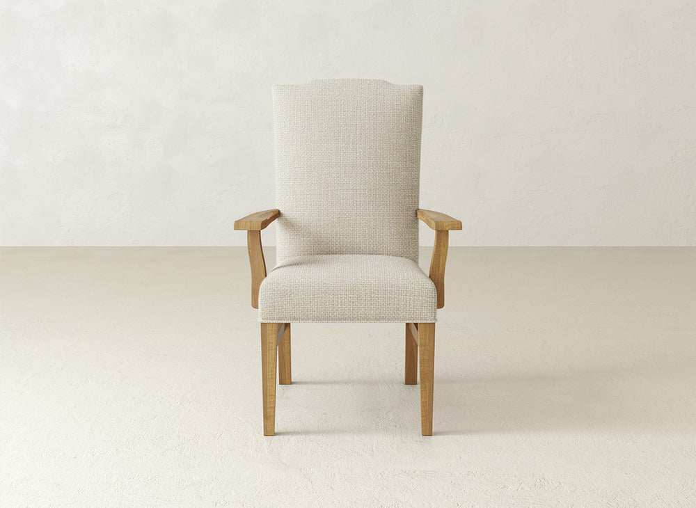 A beige James+James Hadley Arm Dining Chair with wooden legs and armrests is centered in a minimalist room with light textured walls and flooring, combining comfort and elegance as an upholstered dining chair.