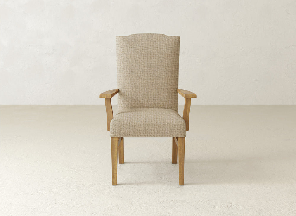 The Hadley Arm Dining Chair by James+James, featuring a beige upholstered seat and backrest, is centered against a plain, light-colored wall and floor.