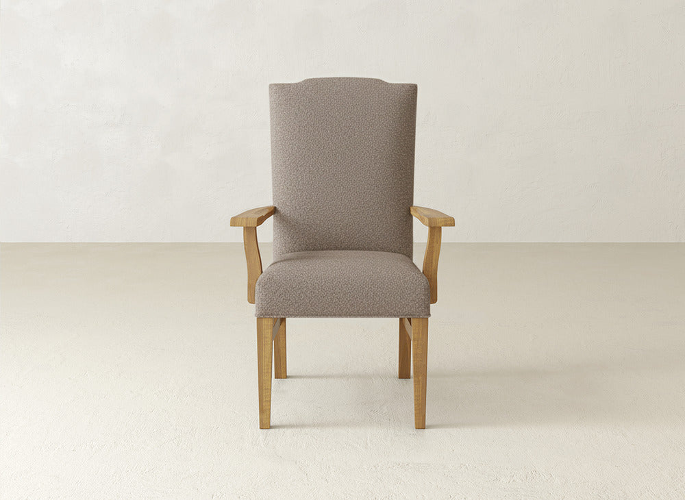 The Hadley Arm Dining Chair by James+James, with a light brown upholstered seat, high backrest, and wooden armrests, sits on a light floor against an off-white wall.