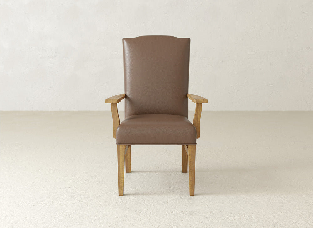The Hadley Arm Dining Chair by James+James, featuring brown fabric upholstery and wooden legs and armrests, is placed on a light floor against a plain beige wall.
