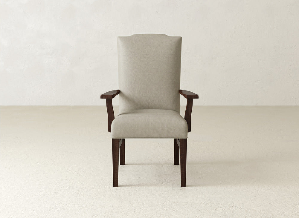 The James+James Hadley Arm Dining Chair is a beige upholstered piece with dark wooden legs and armrests, set on a light textured floor against an off-white wall. It blends classic comfort with timeless style.