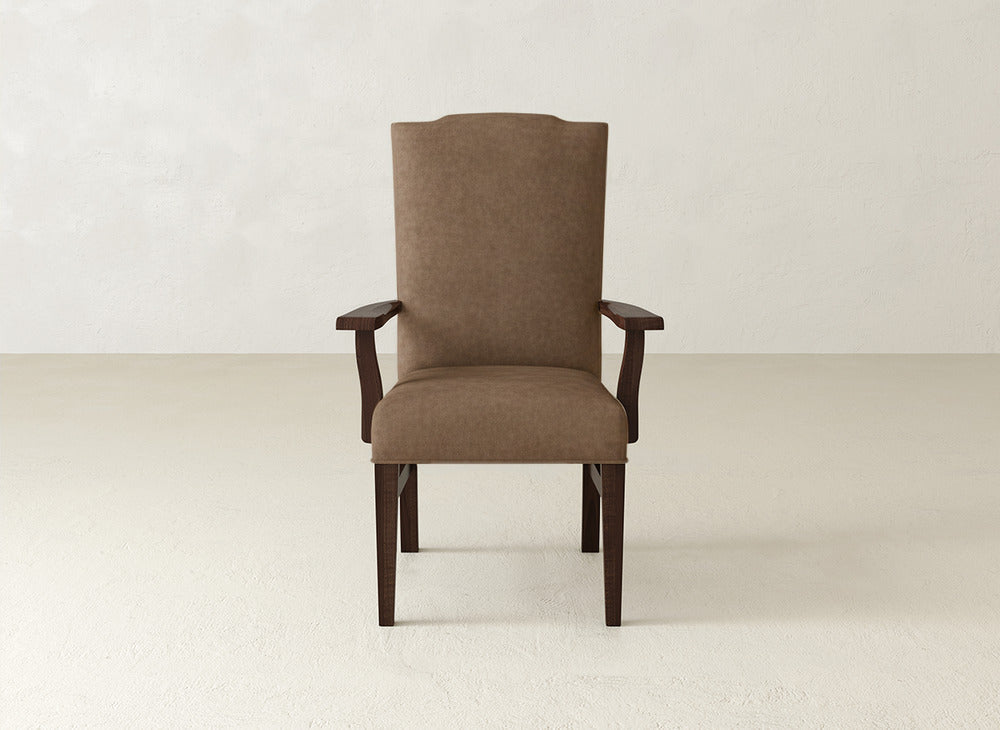 The Hadley Arm Dining Chair by James+James, featuring upholstered seating with wooden armrests and legs, is centered in a minimalist room with a light floor and an off-white wall background.
